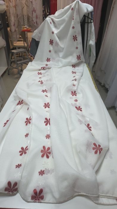 Imaginea prezintă o perdea albă din țesătură ușoară și translucidă, decorată cu modele florale brodate în nuanțe de roșu.
